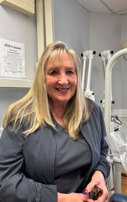 A woman wearing a blue shirt stands behind a dental chair in an office setting, smiling slightly at the camera.